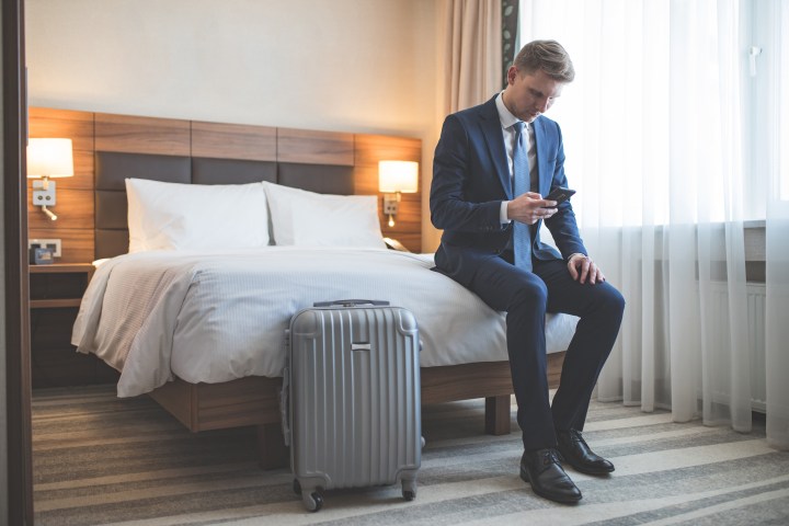 A young man in business formal using his phone while sitting in a hotel room next to a packed suitcase.